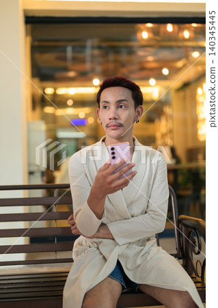 Vertical portrait of non-binary gay Thai man looking away holding smartphone on bench at night 140345445
