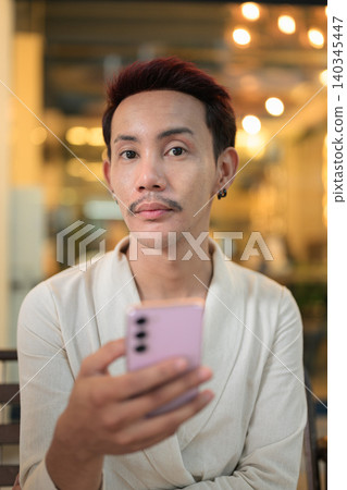 Vertical close up portrait of non-binary gay Thai man looking at camera holding smartphone at night Vertical close up portrait of non-binary gay Thai man looking at camera holding smartphone at night 140345447