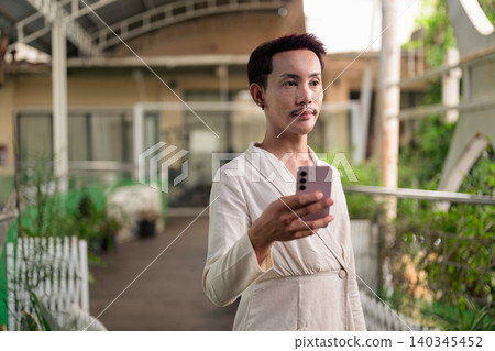 Non-binary gay Thai man looking away holding smartphone on outdoor covered patio walkway Non-binary gay Thai man looking away holding smartphone on outdoor covered patio walkway 140345452