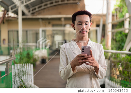 Non-binary gay Thai man typing message on smartphone on outdoor patio walkway Non-binary gay Thai man typing message on smartphone on outdoor patio walkway 140345453