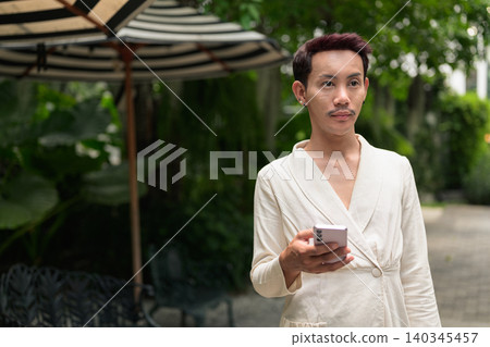 Non-binary gay Thai man looking away while holding smartphone in outdoor garden patio Non-binary gay Thai man looking away while holding smartphone in outdoor garden patio 140345457