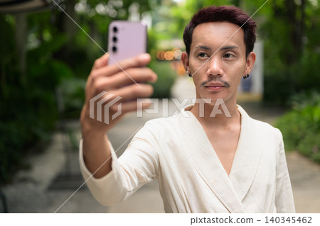 Non-binary gay Thai man looking away while holding smartphone taking selfie in outdoor garden Non-binary gay Thai man looking away while holding smartphone taking selfie in outdoor garden 140345462