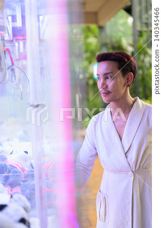 Vertical portrait of non-binary gay Thai man looking at claw crane arcade game machine Vertical portrait of non-binary gay Thai man looking at claw crane arcade game machine 140345466