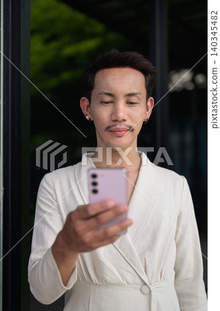 Vertical portrait of non-binary gay Thai man using smartphone outside modern storefront Vertical portrait of non-binary gay Thai man using smartphone outside modern storefront 140345482