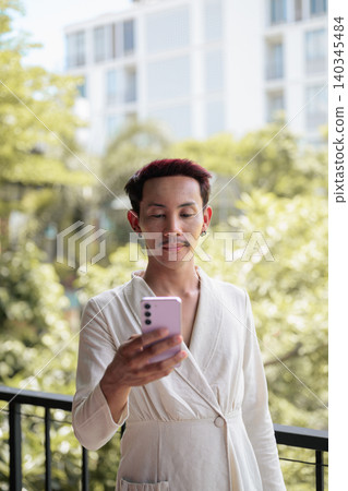 Vertical portrait of non-binary gay Thai man using smartphone on outdoor balcony walkway 140345484