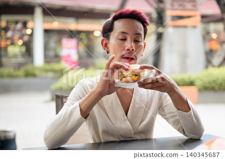 Thai Non-Binary Gay Man Eating Burger at Outdoor Cafe 140345687