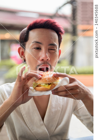 Thai Non-Binary Gay Man Winking While Biting Burger Outdoors Thai Non-Binary Gay Man Winking While Biting Burger Outdoors 140345688