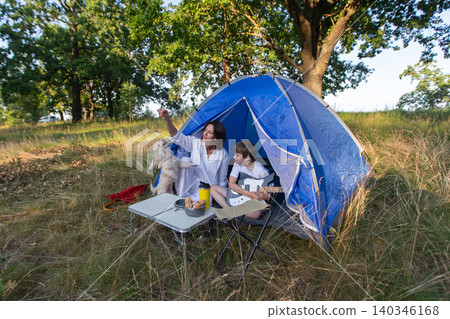 The family enjoys camping in the tent, relaxing in nature and playing music with a dog nearby. 140346168