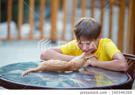 Young boy smiles while playing with an orange cat at a table in a sunny outdoor setting 140346169