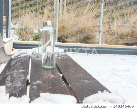 Glass water bottle on snowy wooden bench 140346469