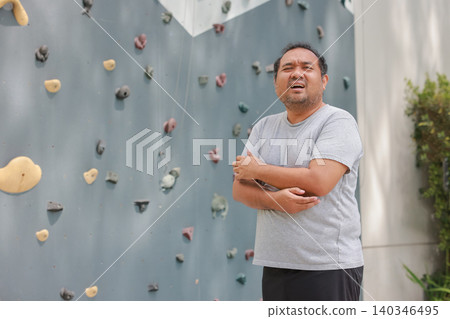 Exhausted overweight Asian man feeling arm muscle pain and holding his elbow after rock climbing workout. Sports injury, physical fatigue, and healthcare concept near outdoor bouldering wall. 140346495