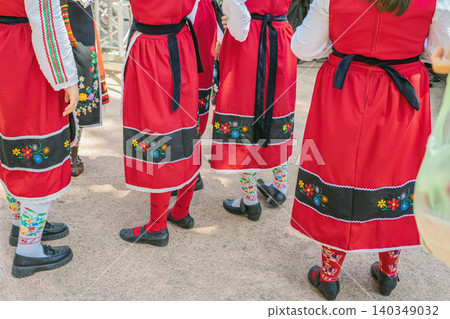 Close view of Bulgarian women wearing red traditional folk costumes with floral embroidery, outdoor cultural event. Heritage clothing, patterned socks, festive folk tradition, ethnic cultural heritage Close view of Bulgarian women wearing red traditional folk costumes with floral embroidery, outdoor cultural event. Heritage clothing, patterned socks, festive folk tradition, ethnic cultural heritage 140349032