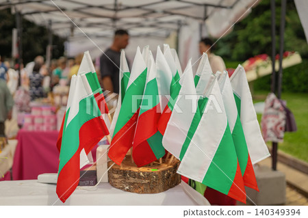 Bulgarian flags stand on market table during Kazanlak Rose Festival with visitors in background. Flag display represents national identity, tourism, celebration, culture, festival merchandise 140349394