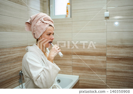 Woman brushing teeth in bathroom wearing bathrobe and towel Woman brushing teeth in bathroom wearing bathrobe and towel 140350908