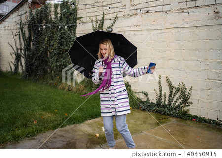 Cheerful young woman dancing in rain holding umbrella and phone Cheerful young woman dancing in rain holding umbrella and phone 140350915