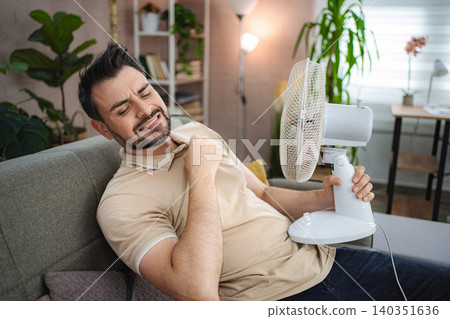 Man suffering from summer heat holding electric fan at home Man suffering from summer heat holding electric fan at home 140351636