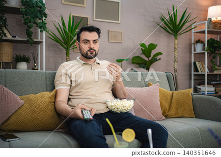 Young man watching television while eating popcorn on the sofa Young man watching television while eating popcorn on the sofa 140351646