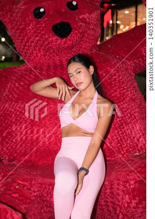 Playful Asian woman in pink sportswear winking next to red teddy bear Playful Asian woman in pink sportswear winking next to red teddy bear 140351901