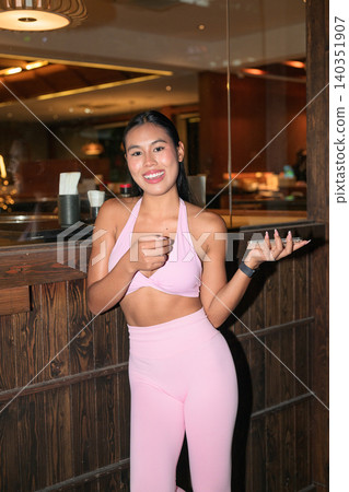 Happy Asian woman in pink fitness wear talking by restaurant window Happy Asian woman in pink fitness wear talking by restaurant window 140351907