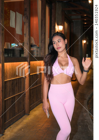 Asian woman in pink fitness clothing walking on night street by restaurant Asian woman in pink fitness clothing walking on night street by restaurant 140351924