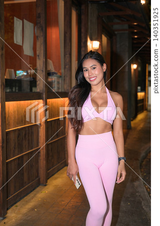 Smiling Asian woman in pink activewear standing outside building at night Smiling Asian woman in pink activewear standing outside building at night 140351925