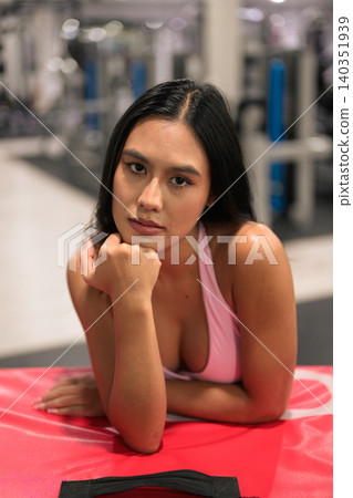 Serious Asian woman in pink activewear leaning on red plyo box in gym Serious Asian woman in pink activewear leaning on red plyo box in gym 140351939