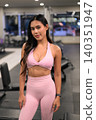 Slightly smiling Asian woman in pink fitness apparel standing in gym 140351947