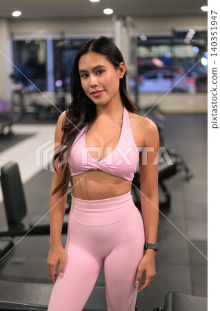 Slightly smiling Asian woman in pink fitness apparel standing in gym Slightly smiling Asian woman in pink fitness apparel standing in gym 140351947