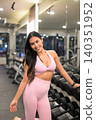 Smiling Asian woman in pink activewear leaning on dumbbell rack in gym 140351952
