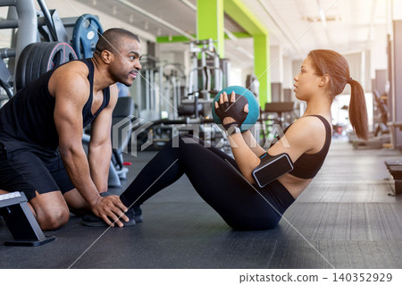 Male trainer assisting woman performing sit-up exercise with medicine ball in gym Male trainer assisting woman performing sit-up exercise with medicine ball in gym 140352929