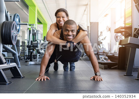 Black man doing push-ups in gym with woman riding on his back Black man doing push-ups in gym with woman riding on his back 140352934
