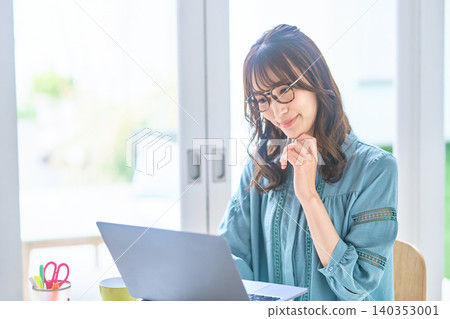 A woman with glasses is thinking while looking at her laptop by the window. 140353001