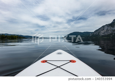 White paddleboard floating on lake Traunsee in the Austrian Alps, capturing a summer recreational moment under a cloudy sky. White paddleboard floating on lake Traunsee in the Austrian Alps, capturing a summer recreational moment under a cloudy sky. 140353994