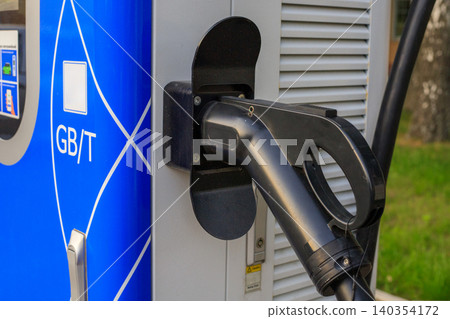 Black EV charger handle is plugged into a blue station terminal side slot. Eco transportation refueling infrastructure. 140354172