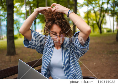 Woman working remotely with laptop in park styling hair Woman working remotely with laptop in park styling hair 140355088