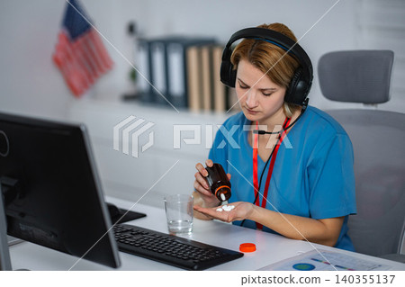 Healthcare worker taking pills at desk wearing headset Healthcare worker taking pills at desk wearing headset 140355137
