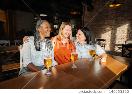 Three diverse women friends enjoying beer and laughing in a pub Three diverse women friends enjoying beer and laughing in a pub 140355230