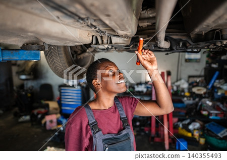 Young female african mechanic inspecting car's undercarriage with flashlight in a garage Young female african mechanic inspecting car's undercarriage with flashlight in a garage 140355493