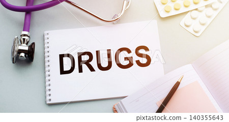 On doctors table is a stethoscope, an open notebook with a black pen and a notepad with word Drugs. On doctors table is a stethoscope, an open notebook with a black pen and a notepad with word Drugs. 140355643