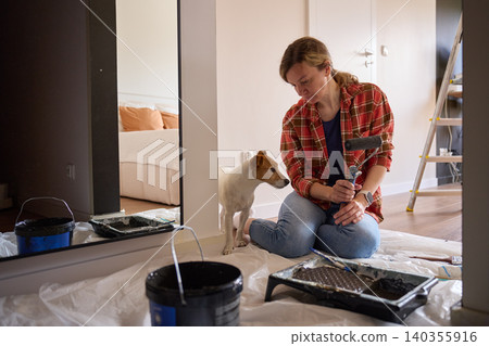 Woman sits on protective sheet holding paint roller beside dog, tray, bucket and ladder Woman sits on protective sheet holding paint roller beside dog, tray, bucket and ladder 140355916