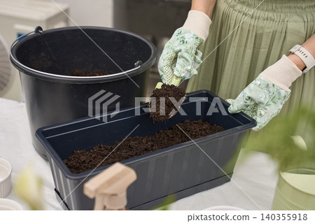 Woman fills black planter box with potting soil using a hand trowel at outdoor garden table 140355918