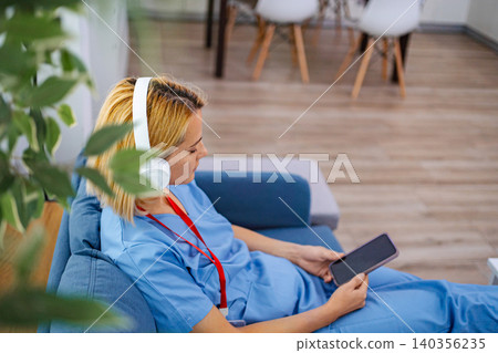 Nurse relaxing on sofa listening to music on headphones and using smartphone Nurse relaxing on sofa listening to music on headphones and using smartphone 140356235