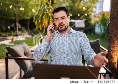 Businessman talking on smartphone feeling frustrated working outdoors Businessman talking on smartphone feeling frustrated working outdoors 140356272