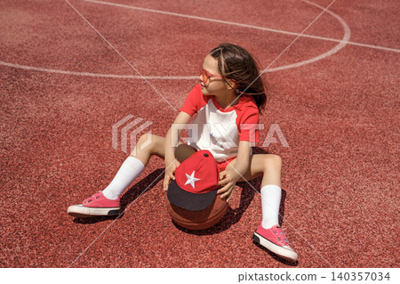 A long-haired girl in sunglasses is relaxing on the rubber surface of the sports field after playing basketball.  A child with a ball 140357034