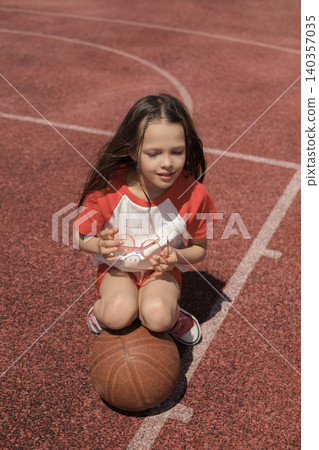 A beautiful girl in sports clothes is sitting with a basketball on the playground with the marking of the playing field A beautiful girl in sports clothes is sitting with a basketball on the playground with the marking of the playing field 140357035