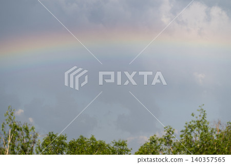 An arch of rainbow soars over vibrant green treetops and peaceful surroundings. Vibrant rainbow stretches across lush treetops as hiker admires breathtaking view with joy 140357565
