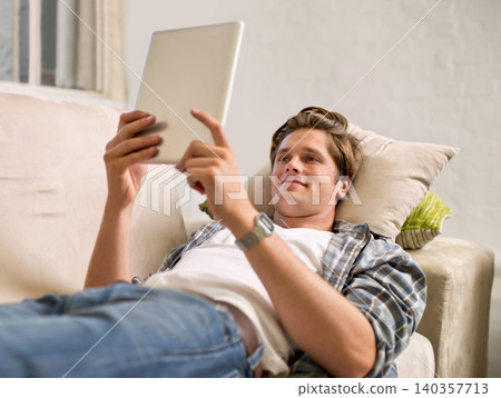 Tablet, relax and man on sofa in home for social media, networking and browse website. Happy, smile and person lying on couch with digital tech for internet, connection and streaming in living room 140357713