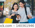 Two women are looking at a cell phone while holding shopping bags 140361946