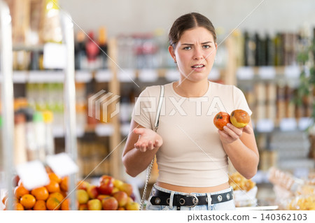 In store, frustrated girl holds rotten fruits in hands. In store, frustrated girl holds rotten fruits in hands. 140362103
