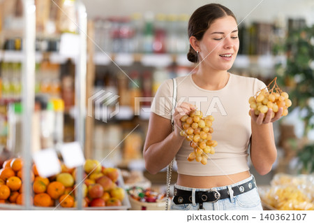 Near counter, young woman client takes out grape from pile of fruits, chooses ripe fruits 140362107